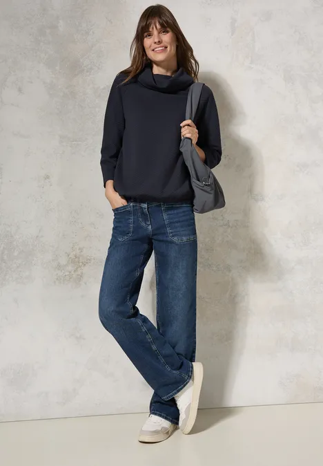 Frau mit dunkelblauem Pullover, Jeans und Tasche lehnt lächelnd an einer Wand.